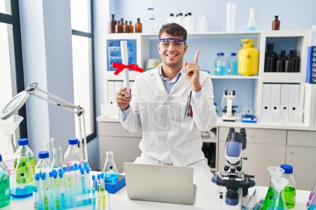 Téléchargez les photos : Jeune homme hispanique travaillant au laboratoire scientifique titulaire d'un diplôme souriant heureux pointant avec la main et le doigt vers le côté - en image libre de droit