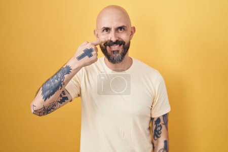 Téléchargez les photos : Homme hispanique avec des tatouages debout sur fond jaune pointant avec le doigt de la main sur le visage et le nez, souriant gai. concept de beauté - en image libre de droit