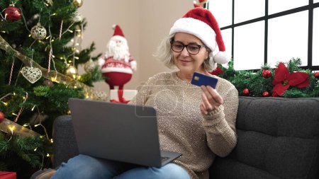Téléchargez les photos : Femme d'âge moyen avec les cheveux gris faire des achats en ligne de Noël avec ordinateur portable à la maison - en image libre de droit