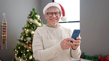 Foto de Hombre de pelo gris de mediana edad usando el teléfono inteligente de pie junto al árbol de Navidad en casa - Imagen libre de derechos