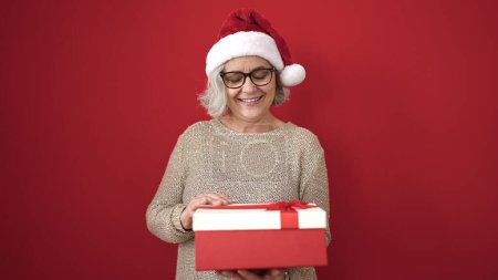 Téléchargez les photos : Femme d'âge moyen avec des cheveux gris portant un chapeau de Noël déballer cadeau sur fond rouge isolé - en image libre de droit