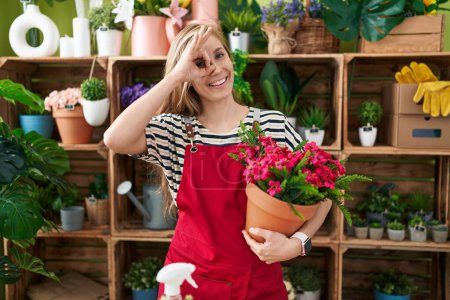 Foto de Joven mujer caucásica trabajando en floristería sosteniendo planta sonriendo feliz haciendo signo de ok con la mano en el ojo mirando a través de los dedos - Imagen libre de derechos