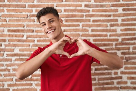 Foto de Joven hombre hispano de pie sobre la pared de ladrillos sonriendo en el amor haciendo forma de símbolo del corazón con las manos. concepto romántico. - Imagen libre de derechos