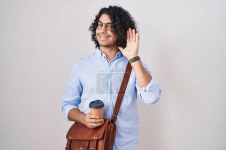 Téléchargez les photos : Homme hispanique aux cheveux bouclés buvant une tasse de café à emporter renonçant à dire bonjour heureux et souriant, geste de bienvenue amical - en image libre de droit