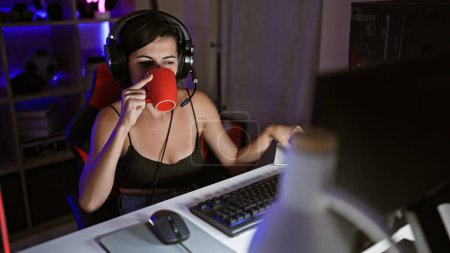 Photo for Radiant young hispanic woman streamer, in her cosy gaming room, playing a futuristic video game, sipping coffee amidst the ambient digital entertainment - Royalty Free Image