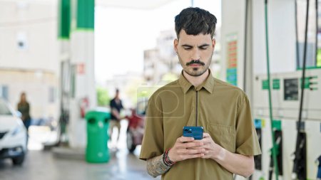 Téléchargez les photos : Jeune homme hispanique utilisant un smartphone avec une expression sérieuse à la station-service - en image libre de droit