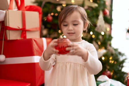 Téléchargez les photos : Adorable fille tenant la décoration de balle debout par arbre de Noël à la maison - en image libre de droit