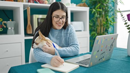 Téléchargez les photos : Jeune femme hispanique avec chihuahua chien en utilisant des notes d'écriture d'ordinateur portable sur ordinateur portable à la salle à manger - en image libre de droit