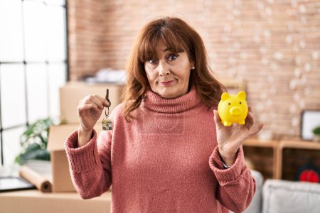 Photo for Middle age hispanic woman holding piggy bank and house keys skeptic and nervous, frowning upset because of problem. negative person. - Royalty Free Image