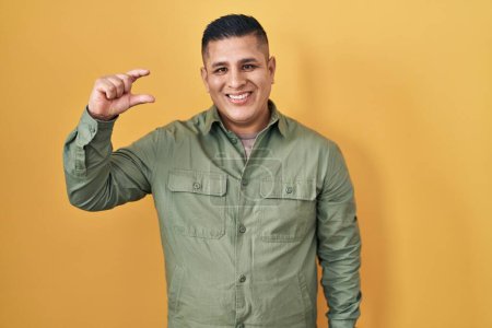 Téléchargez les photos : Jeune homme hispanique debout sur fond jaune geste souriant et confiant avec la main faisant signe de petite taille avec les doigts regardant et la caméra. concept de mesure. - en image libre de droit