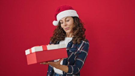 Téléchargez les photos : Moyen Age femme hispanique souriant confiant tenant cadeau de Noël sur fond rouge isolé - en image libre de droit