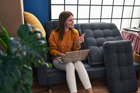 Téléchargez les photos : Jeune femme blonde faisant du shopping avec ordinateur portable et carte de crédit assis sur le canapé à la nouvelle maison - en image libre de droit