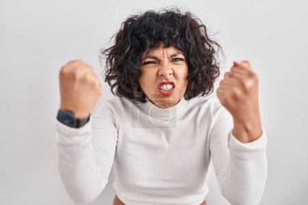 Foto de Mujer hispana con el pelo rizado de pie sobre el fondo aislado enojado y loco levantando los puños frustrado y furioso mientras gritaba con ira. rabia y concepto agresivo. - Imagen libre de derechos