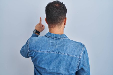Photo for Young hispanic man standing over blue background posing backwards pointing ahead with finger hand - Royalty Free Image