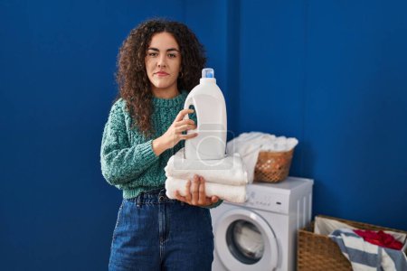 Photo for Young hispanic woman holding laundry and detergent bottle relaxed with serious expression on face. simple and natural looking at the camera. - Royalty Free Image
