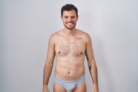 Téléchargez les photos : Jeune homme hispanique debout torse nu portant des sous-vêtements clin d'oeil en regardant la caméra avec une expression sexy, gai et heureux visage. - en image libre de droit
