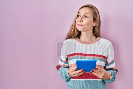 Photo for Young blonde woman holding touchpad smiling looking to the side and staring away thinking. - Royalty Free Image
