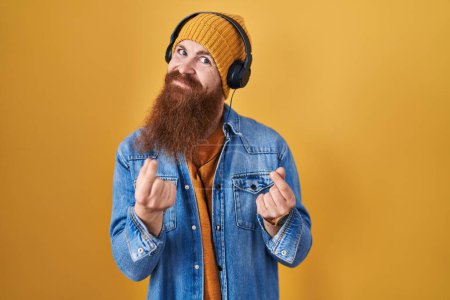 Photo for Caucasian man with long beard listening to music using headphones doing money gesture with hands, asking for salary payment, millionaire business - Royalty Free Image