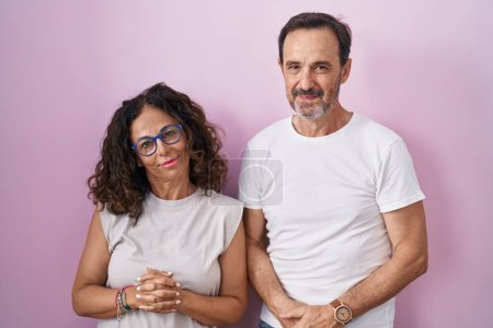 Téléchargez les photos : Couple hispanique du Moyen Âge ensemble sur fond rose avec les mains ensemble et les doigts croisés souriant détendu et joyeux. succès et optimisme - en image libre de droit