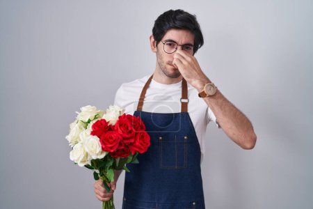 Téléchargez les photos : Jeune homme hispanique tenant un bouquet de roses blanches et rouges sentant quelque chose de puant et dégoûtant, odeur intolérable, retenant le souffle avec les doigts sur le nez. mauvaise odeur - en image libre de droit