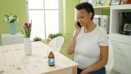 Photo for Young pregnant woman having medical phone consultation sitting on table at dinning room - Royalty Free Image