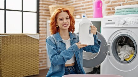 Photo for Young redhead woman washing clothes holding detergent bottle at laundry room - Royalty Free Image