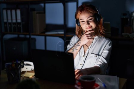 Téléchargez les photos : Jeune femme caucasienne travaillant au bureau la nuit regardant avec confiance la caméra souriant avec les bras croisés et la main levée sur le menton. penser positivement. - en image libre de droit