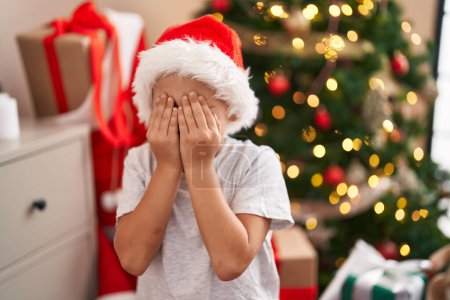 Foto de Adorable niño cubriendo los ojos con las manos de pie junto al árbol de Navidad en casa - Imagen libre de derechos