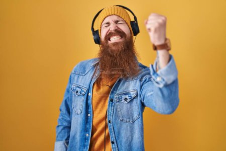 Photo for Caucasian man with long beard listening to music using headphones angry and mad raising fist frustrated and furious while shouting with anger. rage and aggressive concept. - Royalty Free Image