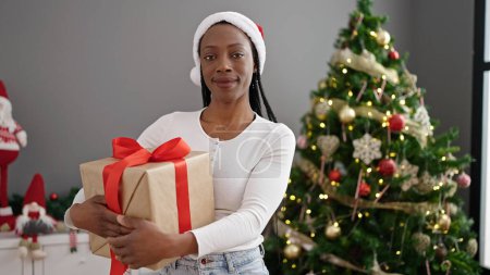 Foto de Mujer afroamericana sosteniendo regalo de pie junto al árbol de Navidad en casa - Imagen libre de derechos