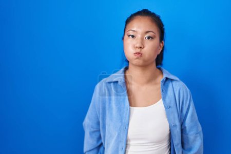 Téléchargez les photos : Asiatique jeune femme debout sur fond bleu joues gonflées avec drôle de visage. bouche gonflée d'air, expression folle. - en image libre de droit