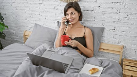Foto de Mujer hispana joven radiante, felizmente charlando por teléfono mientras navega por la computadora portátil, saboreando el café de la mañana en un dormitorio iluminado - Imagen libre de derechos