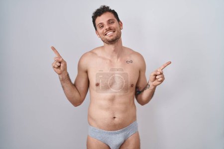Téléchargez les photos : Jeune homme hispanique debout torse nu portant des sous-vêtements souriant confiant pointant avec les doigts vers différentes directions. espace de copie pour la publicité - en image libre de droit