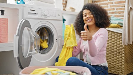 Téléchargez les photos : Afro-américaine femme lavage vêtements faire pouce jusqu'à geste à buanderie - en image libre de droit