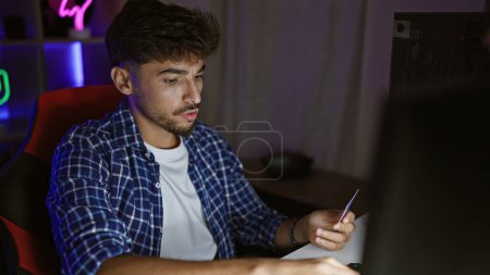 Téléchargez les photos : Flux nocturne passionnant, jeune homme arabe se livre à des jeux virtuels, en utilisant l'ordinateur et la carte de crédit dans la salle de jeux intérieure - en image libre de droit