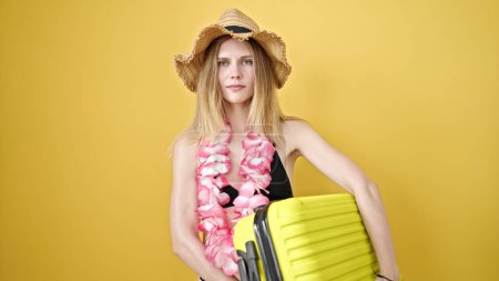 Foto de Joven turista rubia con bikini y sombrero de verano sosteniendo la maleta sobre un fondo amarillo aislado - Imagen libre de derechos