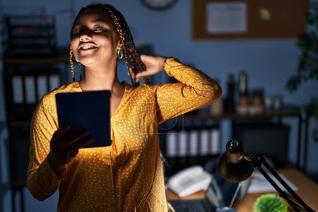 Téléchargez les photos : Femme afro-américaine avec des tresses travaillant au bureau la nuit avec tablette souriant confiant toucher les cheveux avec geste de la main vers le haut, posant attrayant et à la mode - en image libre de droit