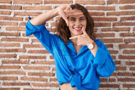 Téléchargez les photos : Belle femme brune debout sur un mur de briques souriant faisant cadre avec les mains et les doigts avec le visage heureux. concept de créativité et photographie. - en image libre de droit