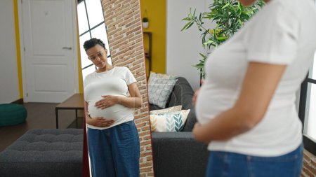 Foto de Mujer embarazada joven mirando vientre en el espejo en casa - Imagen libre de derechos