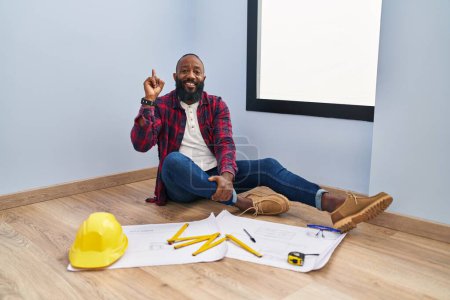 Téléchargez les photos : Afro-Américain assis sur le sol à la nouvelle maison regardant les plans montrant et pointant vers le haut avec le doigt numéro un tout en souriant confiant et heureux. - en image libre de droit