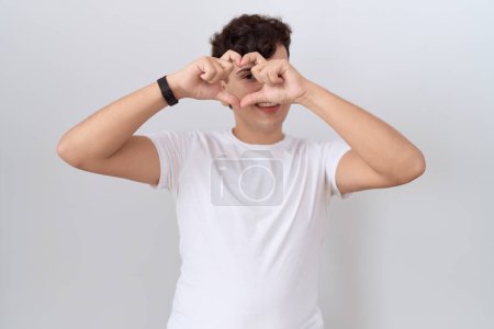 Téléchargez les photos : Jeune homme non binaire portant un t-shirt blanc décontracté en forme de coeur avec la main et les doigts souriant en regardant à travers le signe - en image libre de droit