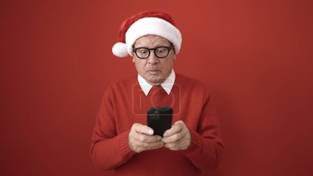Foto de Senior usando sombrero de Navidad usando teléfono inteligente sobre fondo rojo aislado - Imagen libre de derechos