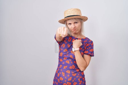 Foto de Mujer caucásica joven con vestido de flores y sombrero de verano puñetazo puño para luchar, ataque agresivo y enojado, amenaza y violencia - Imagen libre de derechos