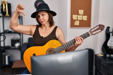 Photo for Middle age hispanic woman playing classic guitar at music studio with angry face, negative sign showing dislike with thumbs down, rejection concept - Royalty Free Image