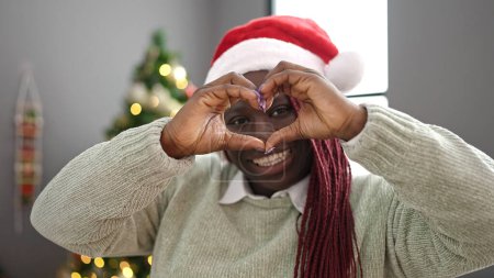 Téléchargez les photos : Femme africaine avec des cheveux tressés souriant par arbre de Noël faisant forme de coeur avec les mains à la maison - en image libre de droit