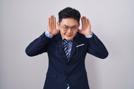 Photo for Young asian man wearing business suit and tie trying to hear both hands on ear gesture, curious for gossip. hearing problem, deaf - Royalty Free Image
