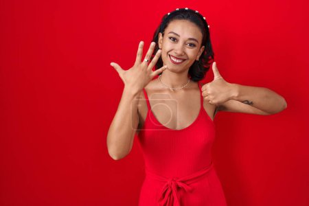 Foto de Mujer hispana joven de pie sobre fondo rojo mostrando y señalando hacia arriba con los dedos número seis mientras sonríe confiado y feliz. - Imagen libre de derechos