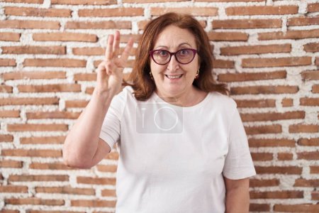 Foto de Mujer mayor con gafas de pie sobre la pared de ladrillos mostrando y apuntando hacia arriba con los dedos número tres mientras sonríe confiado y feliz. - Imagen libre de derechos