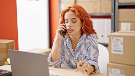 Téléchargez les photos : Jeune rousse femme ecommerce travailleur d'affaires en utilisant un ordinateur portable parlant sur smartphone au bureau - en image libre de droit