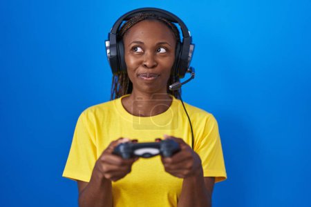 Téléchargez les photos : Femme afro-américaine jouer à des jeux vidéo souriant regardant vers le côté et regardant loin en pensant. - en image libre de droit
